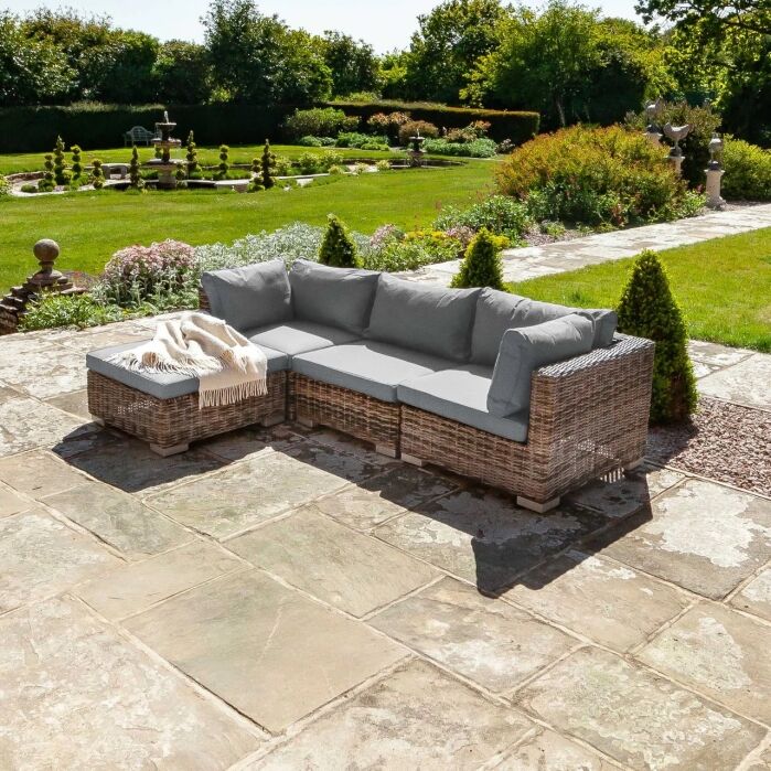 Wicker outdoor sectional sofa with gray cushions and a beige throw on a stone patio, overlooking a landscaped garden with grass, shrubs, and a fountain.