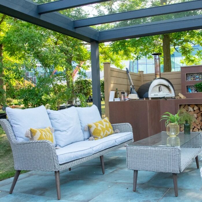 Outdoor patio with a wicker sofa, glass table, wood-fired pizza oven, and stacked firewood, set under a pergola surrounded by trees and greenery.