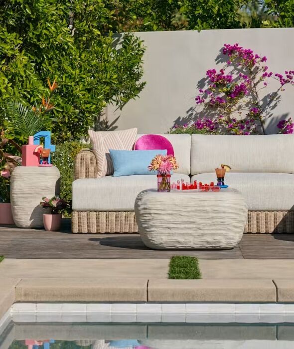 Outdoor patio with a beige wicker sofa, decorative cushions, two side tables with drinks and flowers, and pink flowers climbing a wall, beside a pool.