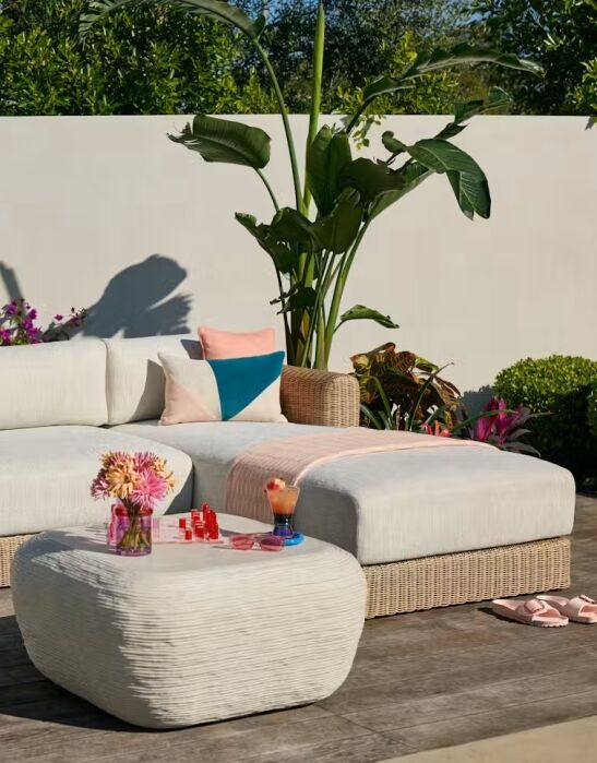 Outdoor patio with a wicker sectional sofa, white cushions, a matching ottoman, tropical plants, flowers, a drink, and a chess set on a wooden deck.