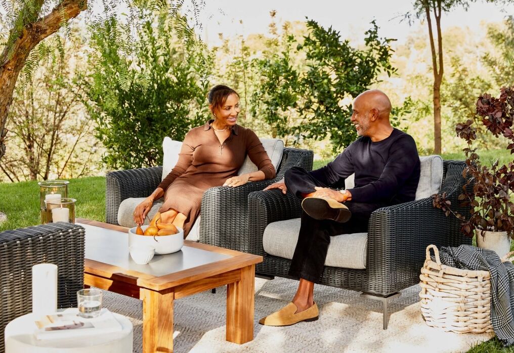 Two people sit on outdoor wicker armchairs, conversing and smiling in a sunny garden setting with a wooden table and greenery in the background.