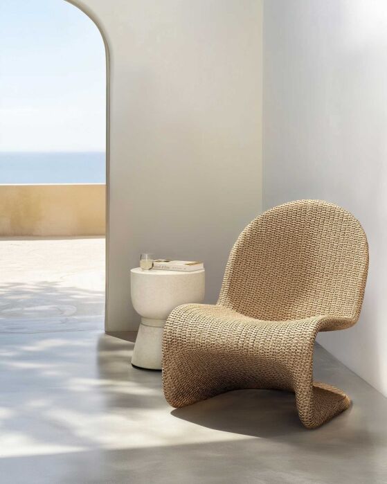 A modern woven lounge chair and small round side table sit in a minimal sunlit room with an arched doorway overlooking a serene outdoor view.