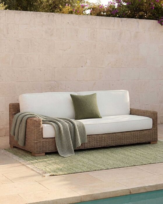 A wicker outdoor sofa with white cushions, a green throw blanket, and a green pillow sits on a green rug beside a swimming pool, against a stone wall.