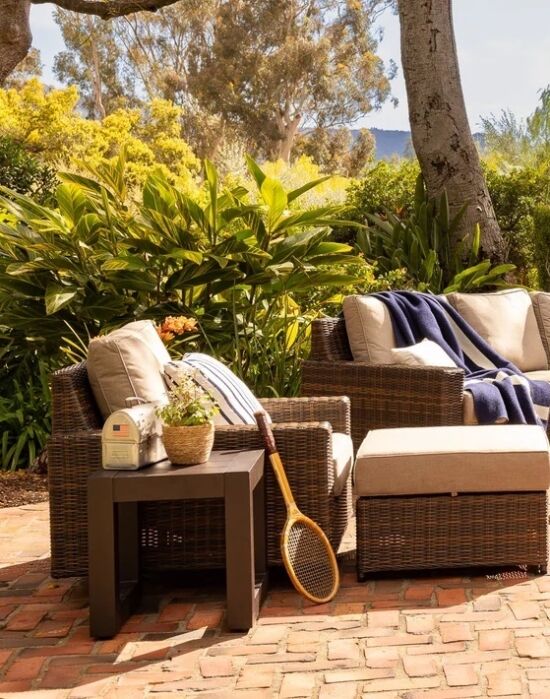 Wicker outdoor seating with cushions, a table holding a potted plant and radio, and a tennis racket on a brick patio surrounded by greenery.