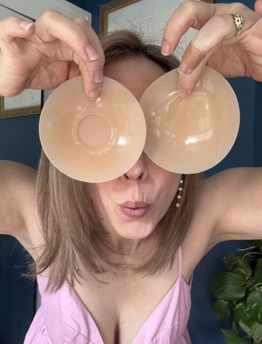 Woman holds two nude-colored silicone bra inserts over her eyes and puckers her lips, posing playfully in a pink top.