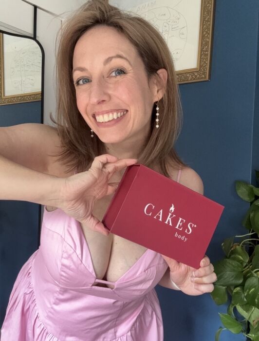 Woman in a pink dress smiling and holding a red box labeled "CAKES body" in front of her, standing indoors next to a plant.