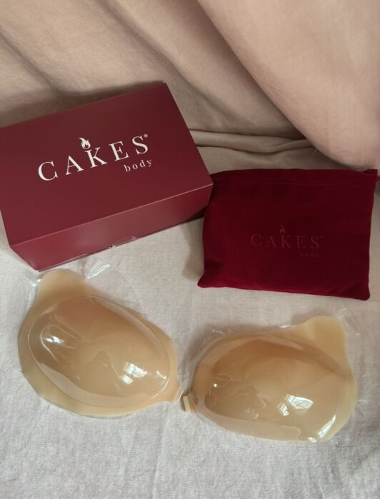 Two adhesive silicone bra inserts are displayed on a surface next to a red CAKES body box and a red storage pouch.