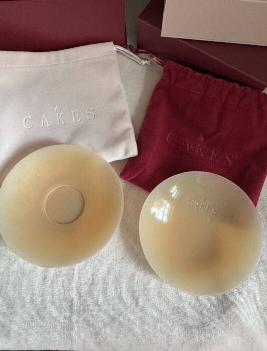 Two round, beige silicone nipple covers are displayed on a white surface, with pink and red CAKES branded drawstring storage bags in the background.