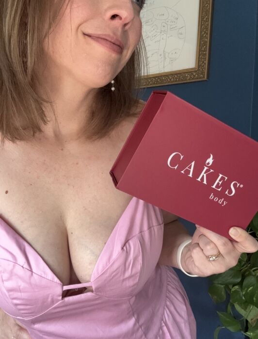A person in a pink top holds a red box labeled "CAKES body." A framed diagram and a plant are visible in the background.
