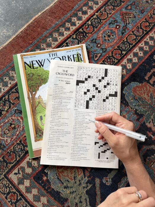 A person completes a crossword puzzle in a New Yorker magazine while seated on a patterned rug; another issue of the magazine is partially visible beneath.