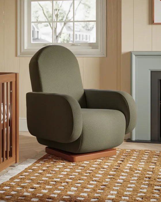 A green upholstered armchair sits on a patterned rug in a cozy room with a window, crib, fireplace, and basket.
