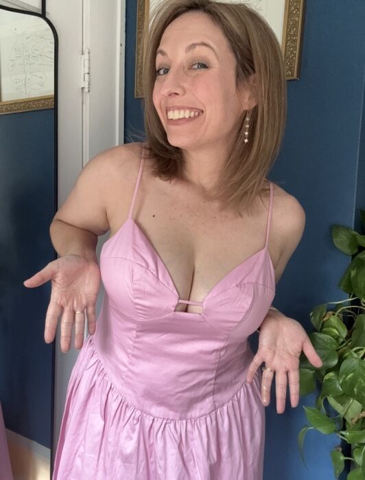 Woman in a pink dress with spaghetti straps stands indoors, smiling and shrugging with palms up.