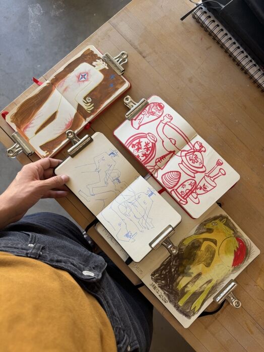 A person holds four sketchbooks open on a table, displaying colorful drawings and sketches, with metal clips holding the pages flat.