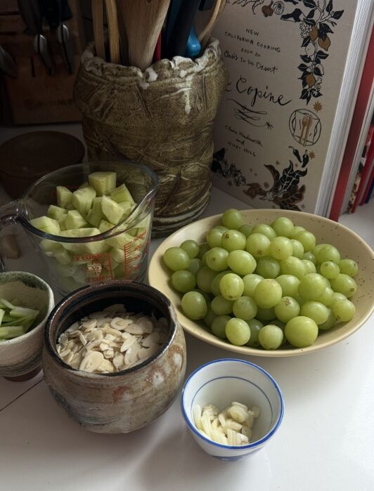 A bowl of green grapes, chopped cucumber, sliced almonds, and garlic are arranged on a counter near a cookbook and utensils.