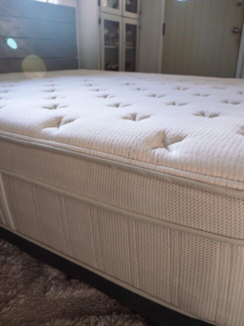 Close-up of a beige mattress with a textured surface and tufted design, resting on a bed frame in a sunlit bedroom.