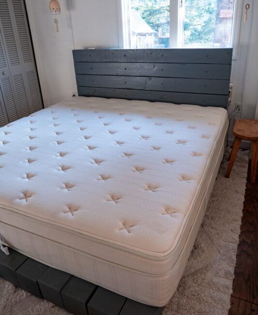 A neatly made bed with a white mattress, wooden headboard, and a small wooden stool in a bright bedroom with windows in the background.