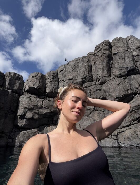 Woman in a black swimsuit stands in front of tall, rocky cliffs under a partly cloudy sky, with water visible at the bottom of the image.