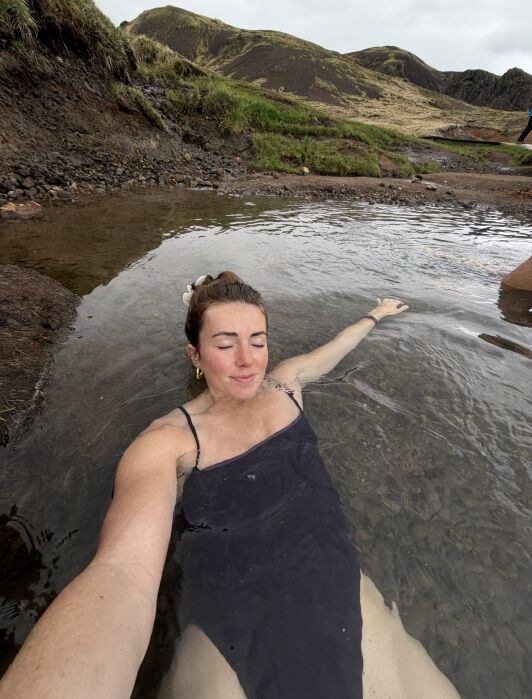 A person relaxes in a shallow natural hot spring surrounded by rocky terrain and grassy hills under a cloudy sky.