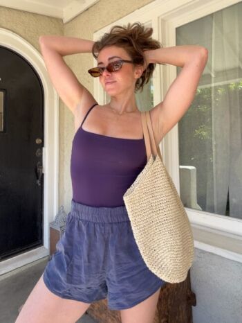 Woman in sunglasses wearing a purple tank top, blue shorts, and a woven tote bag stands with arms raised outside a house.