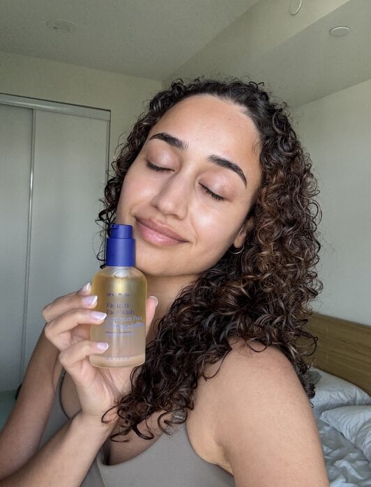 Person with curly hair holds a translucent bottle of skincare product, eyes closed, smiling slightly, standing indoors near a bed and closet.