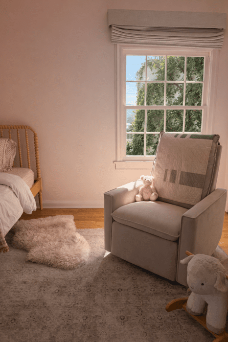 A rocking chair with a pink teddy bear sits by a window in a softly lit child's bedroom, next to a bed, a plush rug, and a toy sheep on the floor.