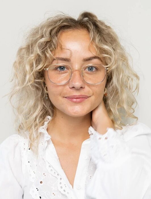 A person with curly blonde hair and glasses, wearing a white shirt, looks at the camera with a neutral expression and one hand touching their neck against a light background.