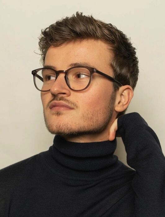 A young man with short brown hair and glasses wears a black turtleneck sweater, looking to the side with his hand resting on his neck against a plain light background.