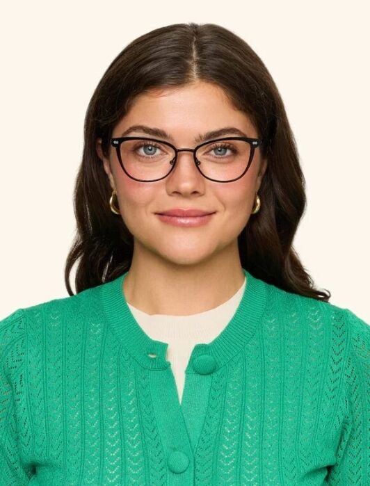 Woman with long brown hair wearing black glasses, a green cardigan, and a white top, standing against a plain light background, looking directly at the camera with a neutral expression.