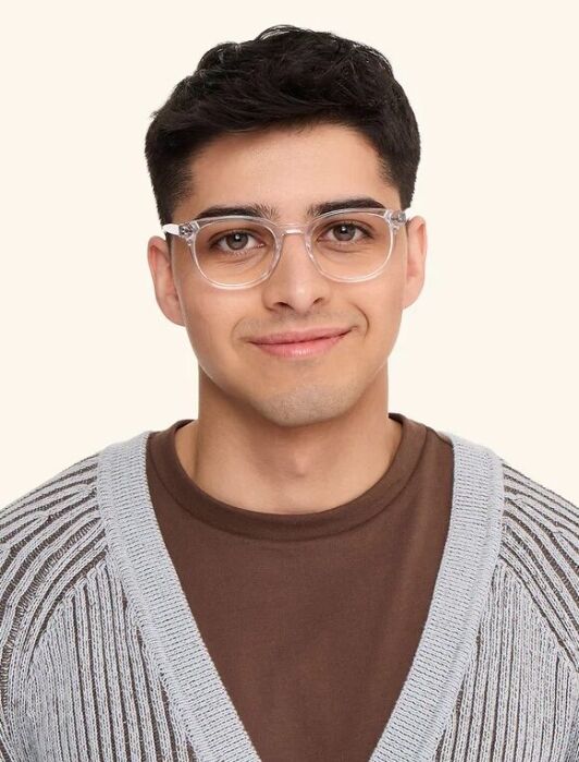 Young man with short dark hair wearing clear eyeglasses, a brown shirt, and a gray knit cardigan, standing against a plain light background.