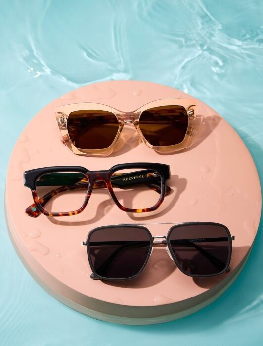 Three pairs of sunglasses—beige, tortoiseshell, and black—are arranged on a round pink platform with water droplets, set against a blue water background.