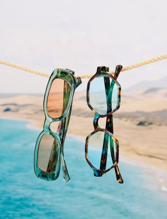 Two pairs of sunglasses hang from a rope above a coastal landscape with blue water and sandy shore in the background.