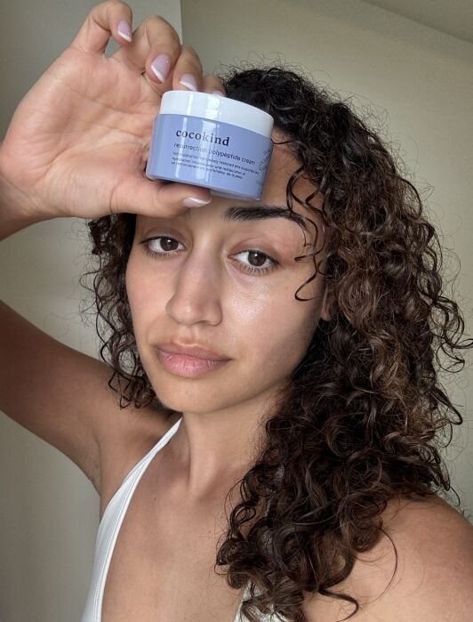 Person with curly hair holds a jar of Cocokind Resurrection Polypeptide Cream next to their face, wearing a white top, in a well-lit indoor setting.