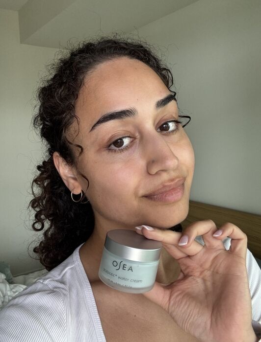 A person with curly hair holds a jar of OSEA Blemish Balm, facing the camera in a well-lit room.