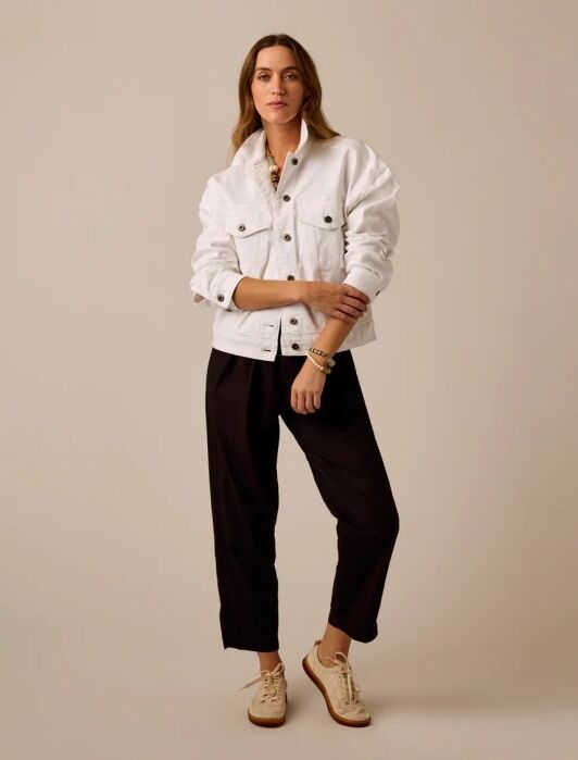 A person stands against a neutral background wearing a white button-up jacket, black pants, and beige lace-up shoes.