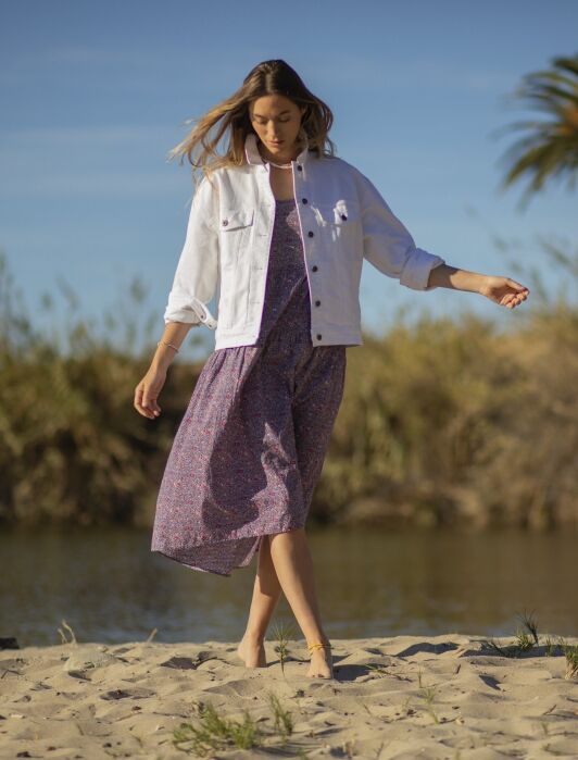 A woman in a white jacket and purple dress walks barefoot on sandy ground near water, with greenery and a palm tree in the background.
