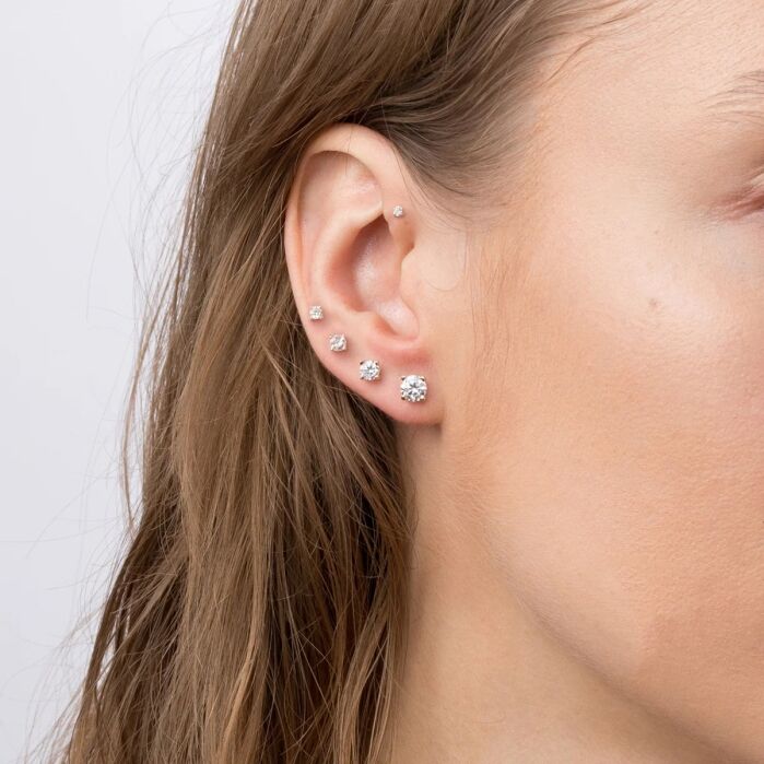 Close-up of a woman's ear with five silver or diamond stud earrings of varying sizes along the ear. Brown hair is tucked behind the ear, and the background is plain white.