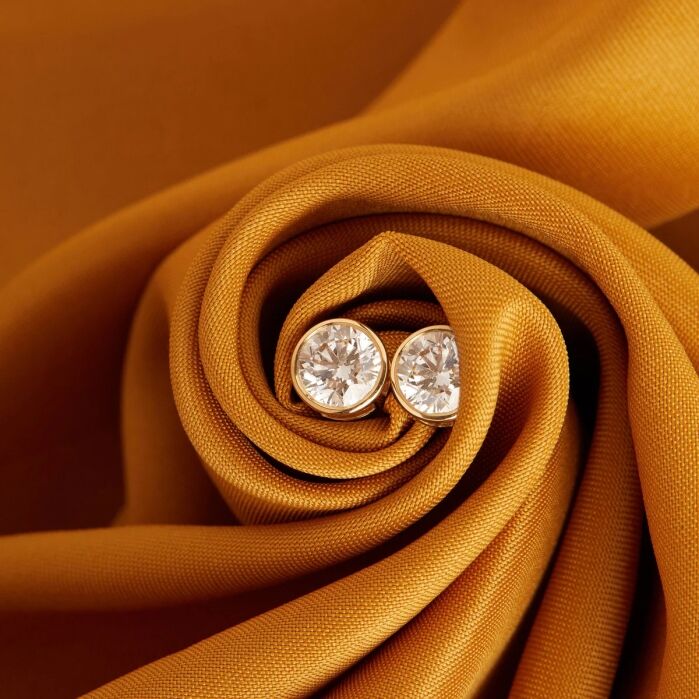 Two round diamond stud earrings set in gold, displayed on a swirl of gold-colored fabric.