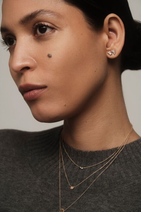 Woman in a grey sweater wears layered gold necklaces and small stud earrings. She has a mole on her left cheek and looks slightly to the side against a neutral background.