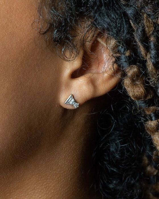 Close-up of a person's ear with a small, silver geometric stud earring, set against a background of curly dark hair with some lighter brown strands.