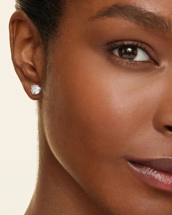Close-up of a woman's face showing her left ear, wearing a round diamond stud earring.