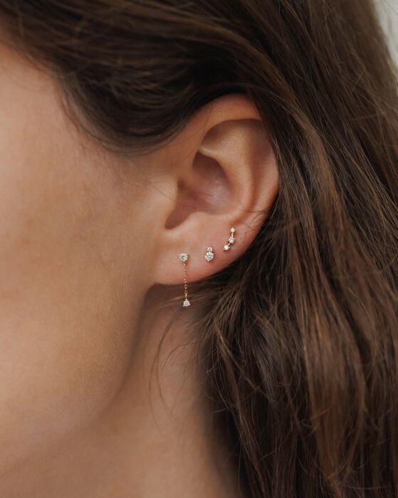 Close-up of a woman's ear with four earrings: a delicate drop earring, two small studs, and a tiny hoop, all in gold, against a background of brown hair.