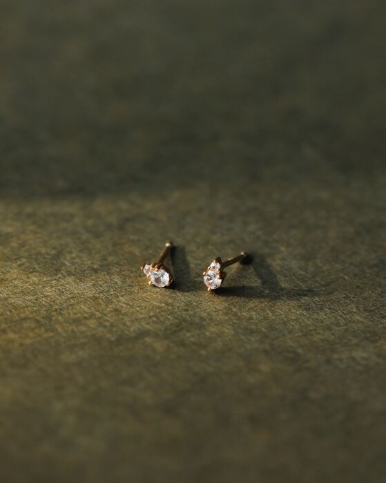 A pair of small gold stud earrings with clear gemstones is displayed on a textured, greenish surface under soft lighting.