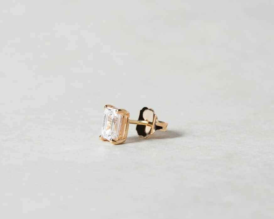A single gold stud earring with a rectangular clear gemstone is shown on a light, neutral background.
