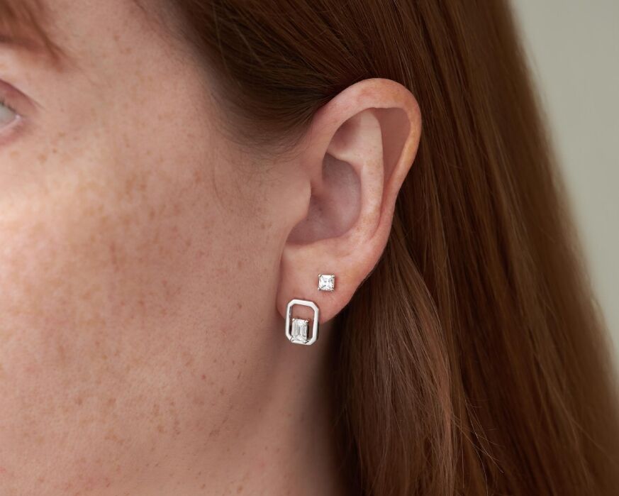 Close-up of a person's ear with two earrings: a small square stud in the upper lobe and a larger rectangular silver earring with an open frame in the lower lobe.
