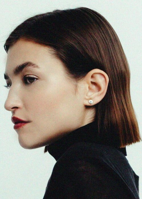 Woman with straight brown bob haircut, wearing a black turtleneck and a round stud earring, shown in profile against a plain light background.