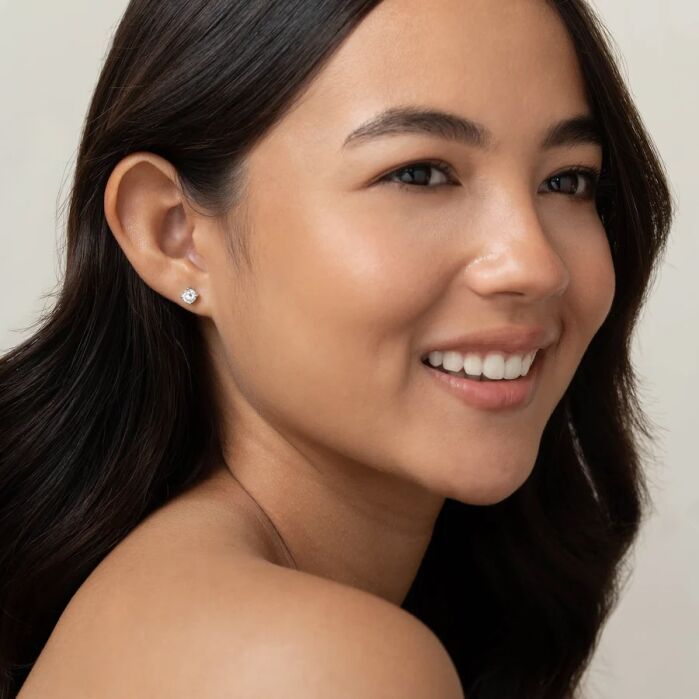 Woman with long dark hair, smiling, wearing a single stud earring, photographed from the shoulders up against a light background.
