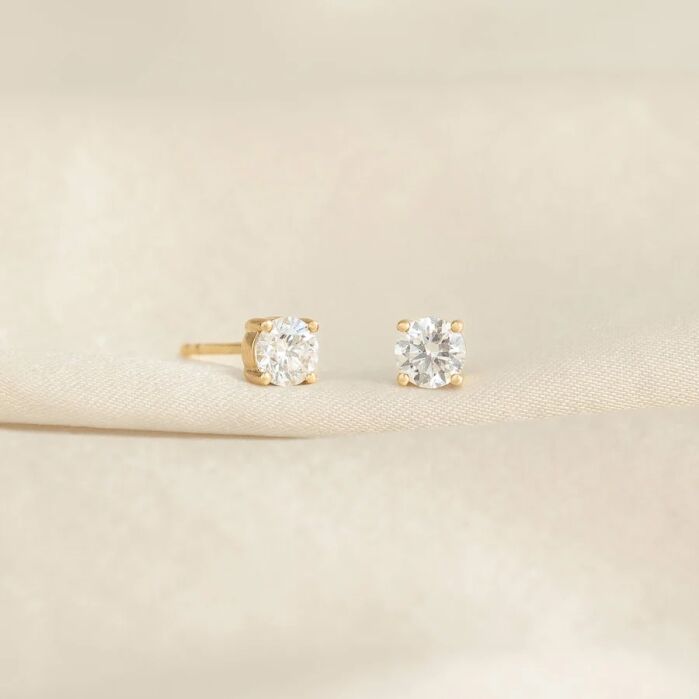 A pair of gold stud earrings with round clear gemstones displayed on a soft beige fabric background.