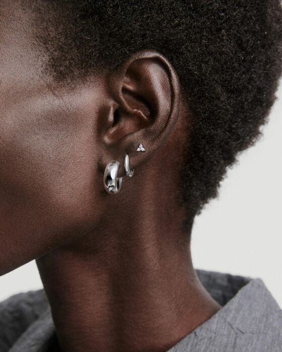Close-up of a person's ear wearing three earrings: a small stud, a medium hoop, and a larger, chunky hoop, with short curly hair visible and part of a gray collared shirt.