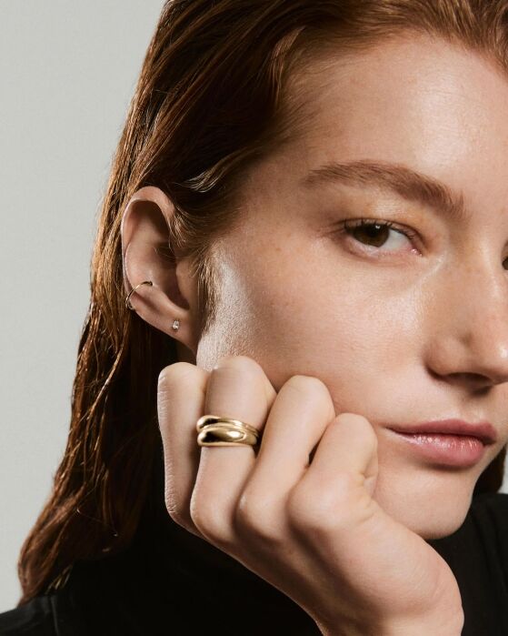 A woman with wet, auburn hair wears a black top, gold ring, and small earrings. Her hand rests on her chin, and she looks slightly past the camera.
