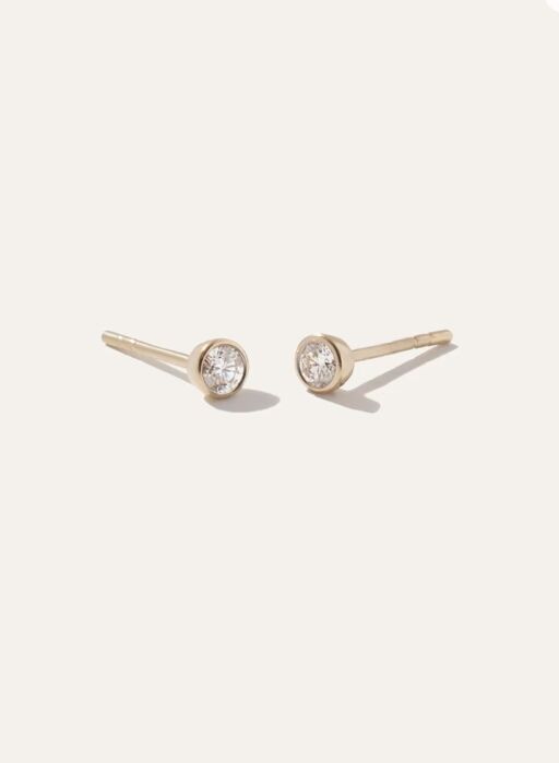 Pair of gold stud earrings each featuring a round clear gemstone set in a simple bezel setting, displayed on a plain light background.
